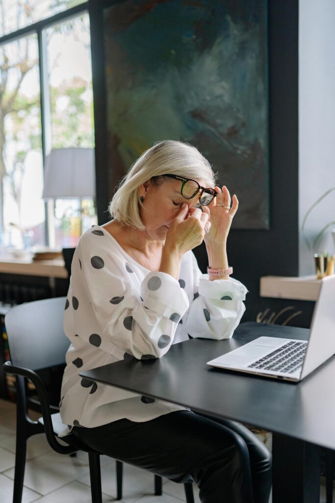Digital Eye Strain. Older woman in a cafe with her laptop, holding her glasses up with one hand and the the bridge of her nose with the other.