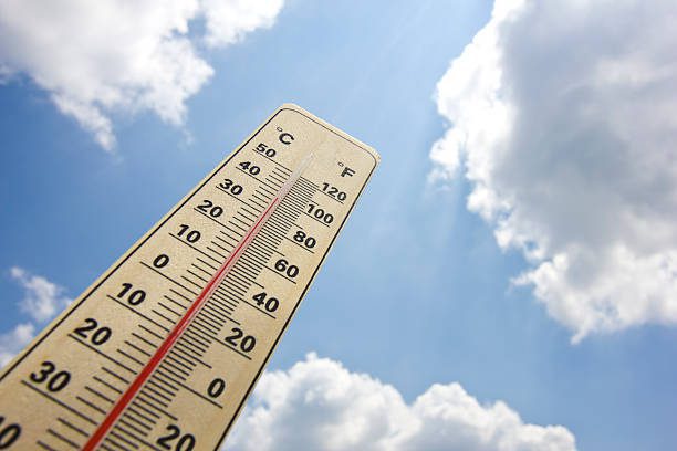 summer heat shown on thermometer with 100 degrees Fahrenheit and 38 degrees Celsius. Sunlight breaking through cumulus clouds on blue sky in background. Photo taken with dslr camera, flash used.