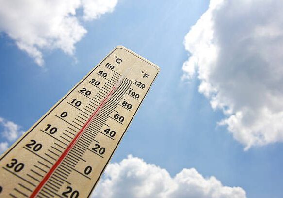 summer heat shown on thermometer with 100 degrees Fahrenheit and 38 degrees Celsius. Sunlight breaking through cumulus clouds on blue sky in background. Photo taken with dslr camera, flash used.